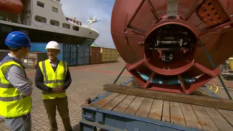 Two men in high-vis are stood quayside. There is the butt of a large buoy next to them on a pallet. A boat is visible in the background.