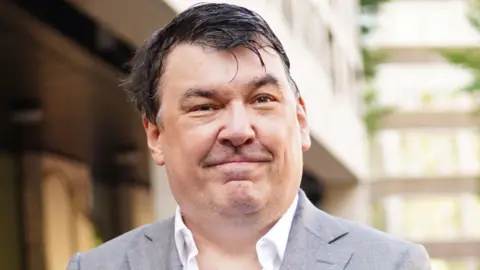 PA Media Graham Linehan in a grey suit and white shirt with no tie posing and looking at the camera outside court