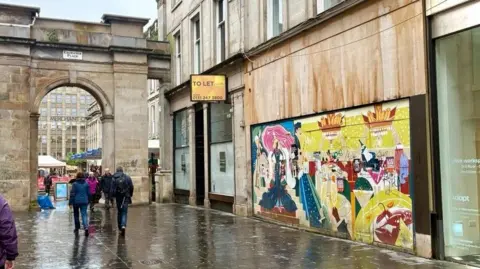 Wide street view of Exchange Place in Glasgow showing a colourful mural covering the boarded frontage of Rogano restaurant, with pedestrians walking past and a stone archway marked Merchant City in the background.