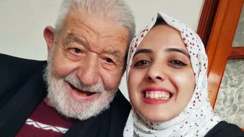 Asmaa Tayeh A selfie of Asmaa, wearing a white headscarf patterned with stars, smiling, wearing winged eyeliner, with her grandad, who has a white beard. He is leaning towards her and his head is touching hers as they look to the camera.