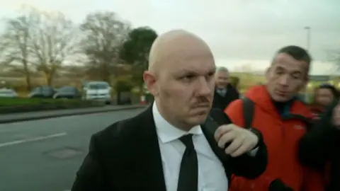 Miles Cross wearing a black blazer and tie. He is straight faced.