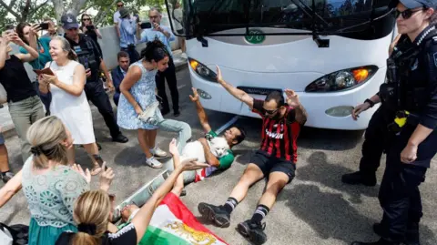 Reuters Two men lie on the ground in front of a coach while police offices and others stand around