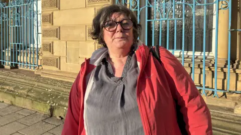 Gemma Dillon/BBC A lady wearing a red jacket and grey top is standing in front of some blue rails. She has brown short hair and glasses.