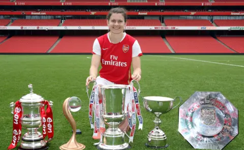 Getty Images Uma jogadora de futebol de cabelos escuros, vestindo um uniforme vermelho e branco do Arsenal, com cinco troféus à sua frente. Ela está ajoelhada em um campo de futebol com assentos vermelhos atrás dela.
