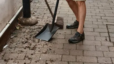 John Whitelegg Pigeon poo on the floor of an alleyway. The bottom of a man's legs can be seen standing next to the droppings. He is also holding a shovel and a brush near to the mess.