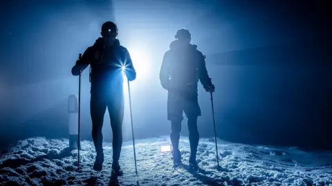 Andrew Hendry / Montane Winter Spine Race Two racers with running poles on snow and silhouetted against a bright back light during nightfall