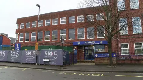 A large brick school building of at least three storeys has a sign saying "St Anne's" and is surrounded by railings