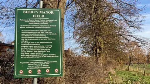 Brian Farmer BBC A green sign saying BUSHY MANOR FIELD. The sign lists a number of regulations people are asked to observe. In the background is a hedge, trees and grass 