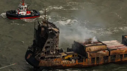 DAN KITWOOD/POOL/EPA-EFE/REX/Shutterstock Smoke billowing from the fire-damaged Solong cargo vessel in the sea. A small tug boat is nearby on the top left.