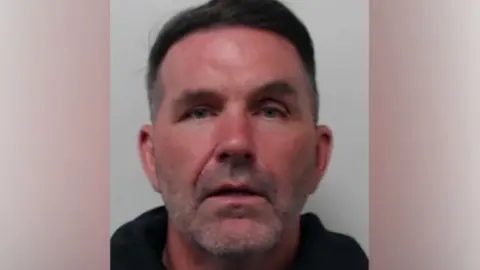 A mugshot of a man with short dark hair wearing a dark top.