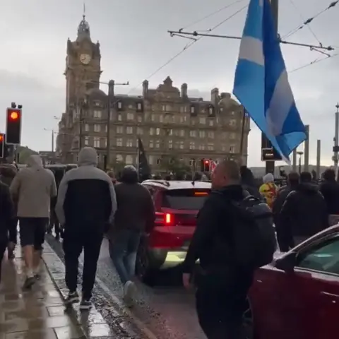 A group of protestors walking down a road among cars. a Saltire is being held up