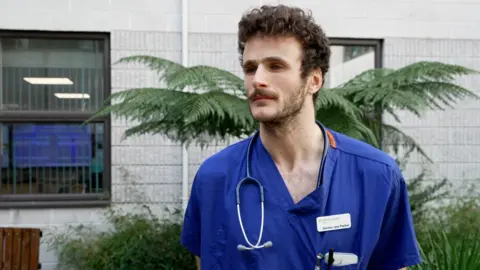 Dr Joe Parker is standing in the healing garden in his blue scrubs with a stethoscope around his neck and an identity badge pinned to his top. He has dark curly hair and a moustache and beard. Behind him is a palm tree, windows and a stone wall.