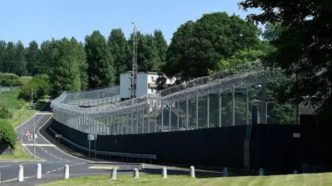 Michael Cooper The perimeter fencing at Hydebank with a road running alongside it with trees throughout the site and a compound behind the wire 