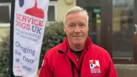 A man with white hair wearing a red jacket. He is looking into the camera and smiling. Behind him to his left is a white banner