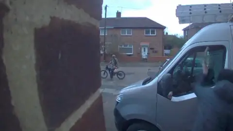 Handout An image from a doorbell camera shows a person is sitting on a dirt bike on a road in the background while in the foreground somebody else hitting the passenger side window of a parked silver van. The attacker is using a weapon, believed to be an axe. It is happening in a housing estate with two-storey red-brick houses visible on the other side of the road.