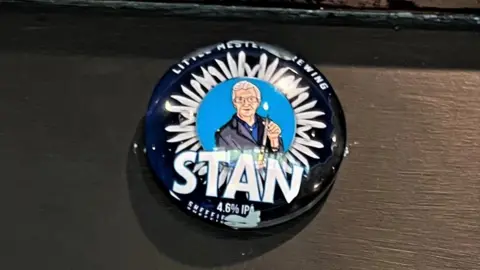 Little Mesters Brewery A circular blue badge depicting a cartoon image of a man with grey hair and glasses, Stan Shaw, holding a knife with a circle of assorted blades around him