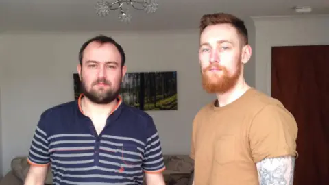 Two men stood in a living room. The man on the left has black hair, black beard, navy polo t-shirt with orange trim and grey stripes. The man on the right has red hair, a red beard, a light brown t-shirt and a heavily tattooed left arm. The living room is cream, with a cream settee, woodland photograph, dark wooden door and a flowery light fitting.