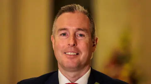 Paul Givan inside Stormont, he is wearing a navy suit and a white shirt with a red tie. He has short white hair.