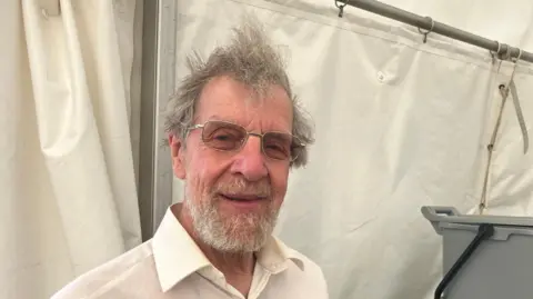 Vikki Irwin/BBC A man with grey hair, some of which sticks upwards, smiles at the camera. He has a thick grey beard. He is wearing a white shirt and is standing inside a white tent. 