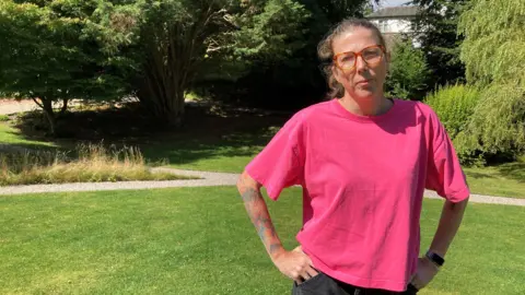 Sarah Wilson is wearing a pink T-shirt and standing in the foreground. She has her hands on her hips and has short, tied back hair and is wearing glasses. In the background there are green trees and she is on green grass.