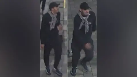 British Transport Police A man wearing a scarfe in a black cap and black top and trousers. He has black trainers with a lighter coloured strip on