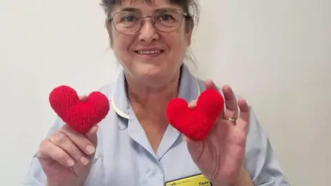 Bury Hospice A woman with brown hair cut into a fringe and glasses, and wearing a light blue hospice uniform with a name badge reading 'Hello my name is Paula' holds up two small knitted red love-hearts to the camera, one in each hand. 