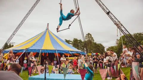 World Jungle An open-air aerial display with a woman hanging from a rope doing the splits in mid-air. She is wearing a bright turquoise leotard. In the background are spectators, some sitting on the grass and some standing and clapping behind. There is also a blue and yellow striped big top and some large mature trees. 