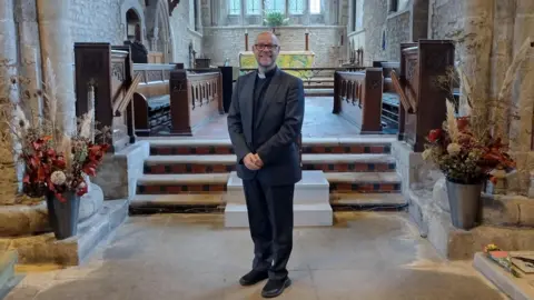 BBC Reverend Canon Martin, in a black suit, standing in a church.