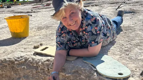Bournemouth University Sandy Toksvig lays on the chalky ground next to a trench and a yellow bucket at an archaeological dig. She is leaning on her elbows and smiling at the camera. She has blonde short hair and is wearing a sunhat, patterned shirt and jeans.