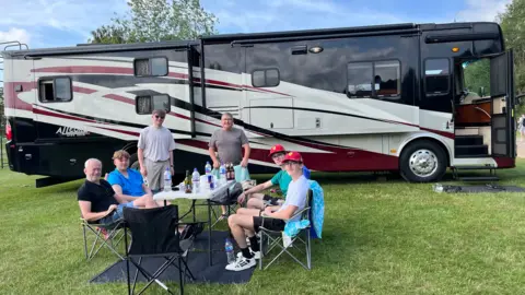 Tom Williams/BBC A group of people sat at a picnic table beside a large RV that is parked on a field.
