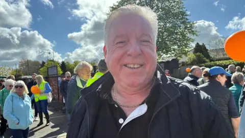 A man with short white hair, a chain around his neck, a black shirt and dark blue jacket, smiles as he stands in the middle of a crowd of people protesting.