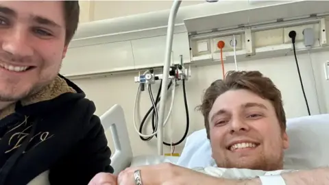 Zak Nelson A smiling man in a hospital bed, his hand raised showing a silver ring on his finger, with a smiling man in a black top sitting by his bedside. 