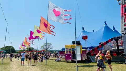 Wight Life A row of fluttering colourful banners arc through this picture of the Isle of Wight Music festival. A big blue tent can be seen to the right with a fairground ride in front of it. Festival goers wander through the shot almost exclusively dressed in shorts under a cloudless blue sky.