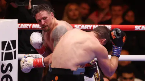Reuters/ Amr Abdallah Dalsh Oleksandr Usyk is wearing white gloves and is facing the camera, having thrown a left hook at Rico Verhoeven, during their WBC Heavyweight title fight in Egypt. Verhoeven has his back to the camera and is ducking away from the punch