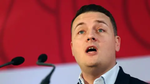 Health Secretary Wes Streeting speaks in front of a microphone. He is standing in front of a red-and-white background.