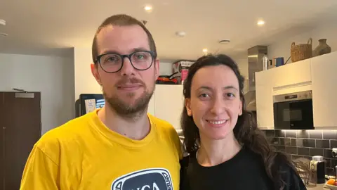 A man in a yellow top stands next to a woman in a black one. He has short brown hair and glasses. She has long dark brown hair