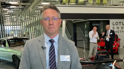 Alex Dunlop/BBC Matt Sanger standing in a room in Lotus. There are Lotus sports cars around him. He is looking directly at the camera and is wearing a pale blue shirt, grey blazer and navy and red patterned tie. He is always wearing a pair of circular glasses.