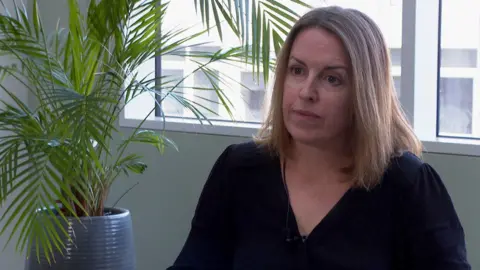 Tessa Gregory has blonde hair and is wearing a dark v-neck top. She's sitting in an office with a houseplant next to her and a window behind her. 