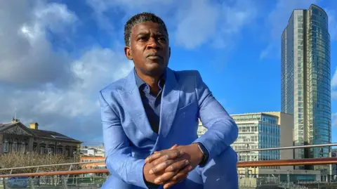A black man pictured wearing a sky blue suit and trousers, he has his hands closed and is wearing a black apple watch. 
