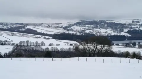 Weather Watchers/SheafofWheat A serene winter landscape dominated by snow-covered fields and rolling hills. In the foreground, there is a large expanse of untouched snow bordered by a simple wire fence. Behind the fence, clusters of leafless trees stand scattered across the terrain, their dark branches contrasting sharply against the white snow.
Further into the background, the hills rise gently, layered with snow and divided by dark lines of hedgerows or stone walls, creating a patchwork effect. A body of water, likely a lake or reservoir, is partially visible among the trees in the mid-ground, adding depth to the scene. The hills in the distance are dotted with more trees and appear to have a mix of open fields and wooded areas.