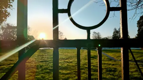The sun streaming through a metal gate in Oxford, with sky as the background to the top half of the picture, and grass the bottom half