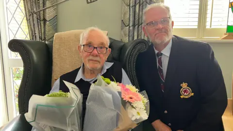 Two men, one sitting in a chair and one kneeling next to it. The man in the chair, Mr Kennett, has short white hair and black glasses. He is wearing a light blue shirt and black tank top and is holding two bunches of flowers. The man next to him also has short white hair and glasses. He is wearing a black jacket with a military patch on its breast, a white shirt and blue, maroon and white tie.