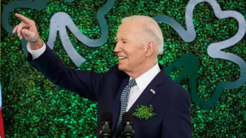 Getty Images A man with white hair in front of a green background wearing a navy suit with white shirt and green tie, he is pointing