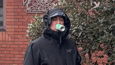 PA Man in black rain coat with hood up walks past red brick court building. A green can is held in his mouth, without his hands holding it.