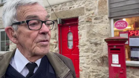 Mike Trenwith is standing outside a stone building which is the Post Office and it has a red door. He has grey hair and wears glasses, a jacket, jumper, white shirt and black tie. There is a post box behind him set in front of a window that has a sign reading POST OFFICE. 