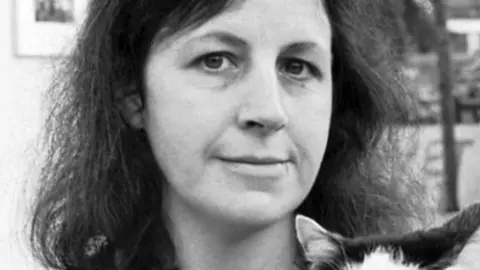 Black-and-white photograph of a woman holding a black-and-white cat. She faces the camera with a calm expression, and the background shows a softly focused indoor gallery or museum space. You can only see the cat's ears.