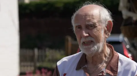 A man with short white hair and a matching beard stands in the driveway of a property with a black car behind him. He is wearing a brown and white shirt. The sun is shining on the side of his face.