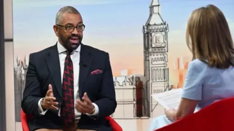 BBC Sir Cleverly is pictured in the Sunday with Laura Kuenssberg studio. He and Victoria Derbyshire, standing in for Laura Kuenssberg, are sitting in red bucket chairs, facing each other. Cleverly wears pale brown trousers, a navy blazer, a white shirt and a blue and claret zigzag stripe tie. Derbyshire is wearing a pale blue dress with short sleeves and has shoulder length, dark blonde hair.