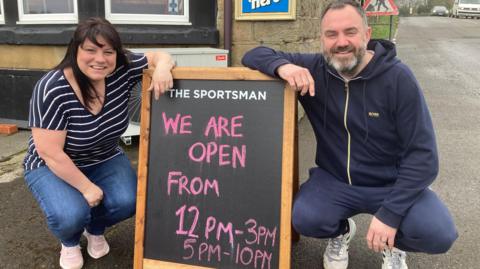 A woman in a stripy t shirt and a man in a blue hoodie squat down next to a chalkboard sign outside a pub.