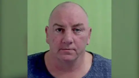 John Meadows a bald middle-aged man, stares directly at the camera in a police mugshot with a green background.